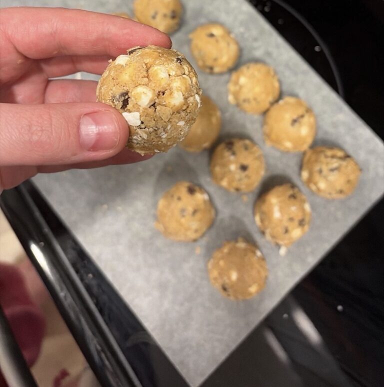 Hand holding a s'mores protein ball with chocolate chips and mini marshmallows over a tray of protein balls