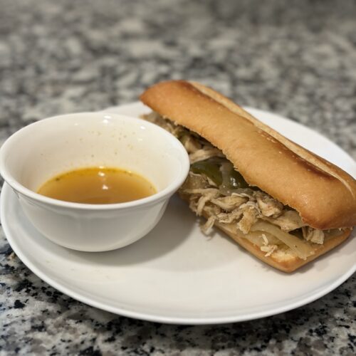 Slow cooker chicken Philly cheesesteak sandwich on a plate with a bowl of dipping juice